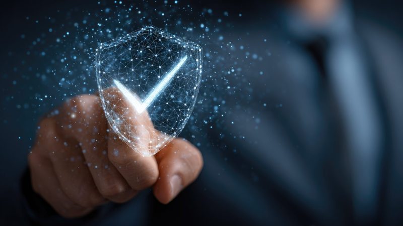 A mans hand interacts with a futuristic holographic shield interface with a checkmark, symbolizing successful protection, security, and data safety in the digital world of technology.